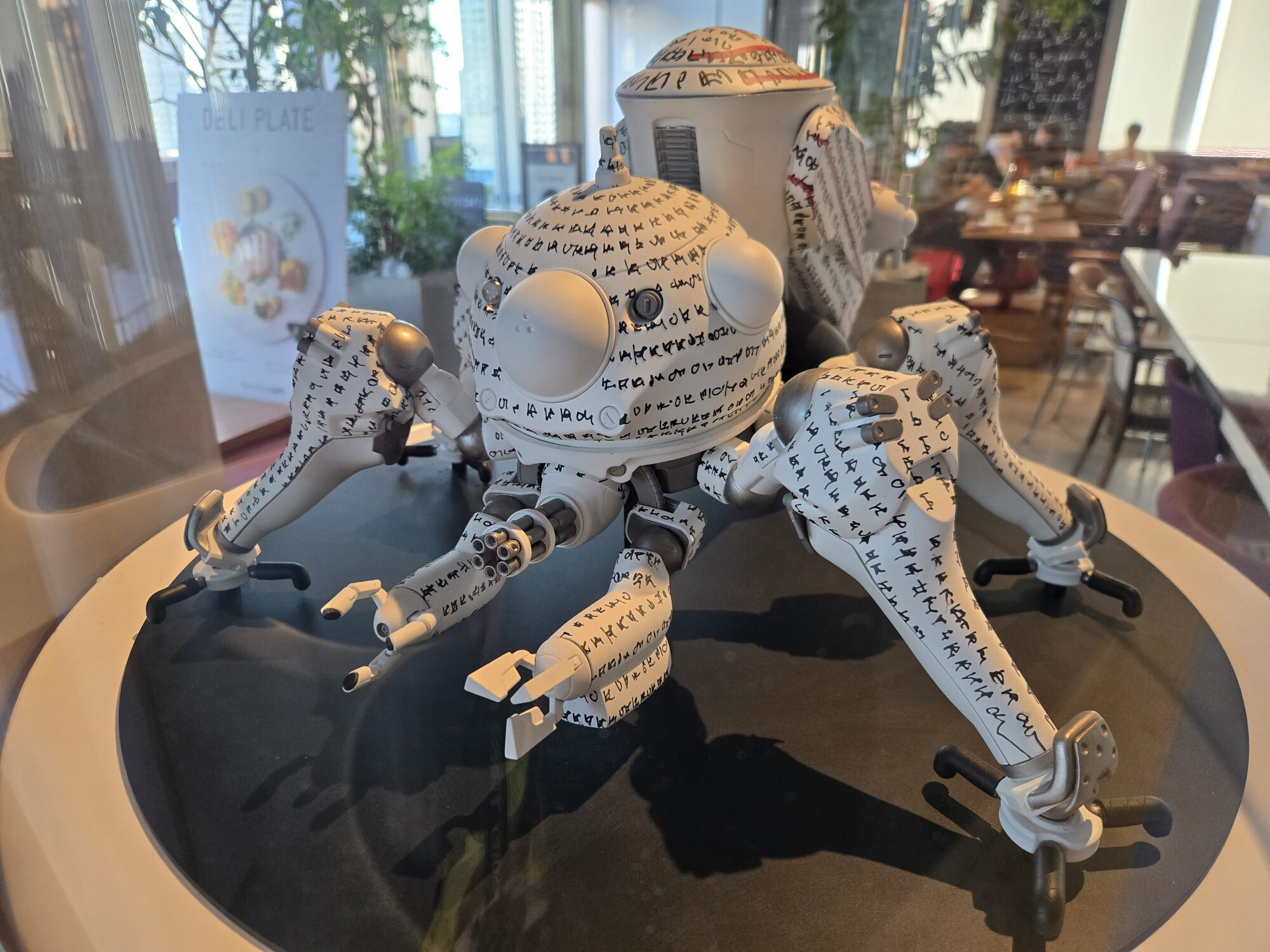 White Tachikoma covered in handwritten kanji text in glass display case at Ghost in the Shell Exhibition café