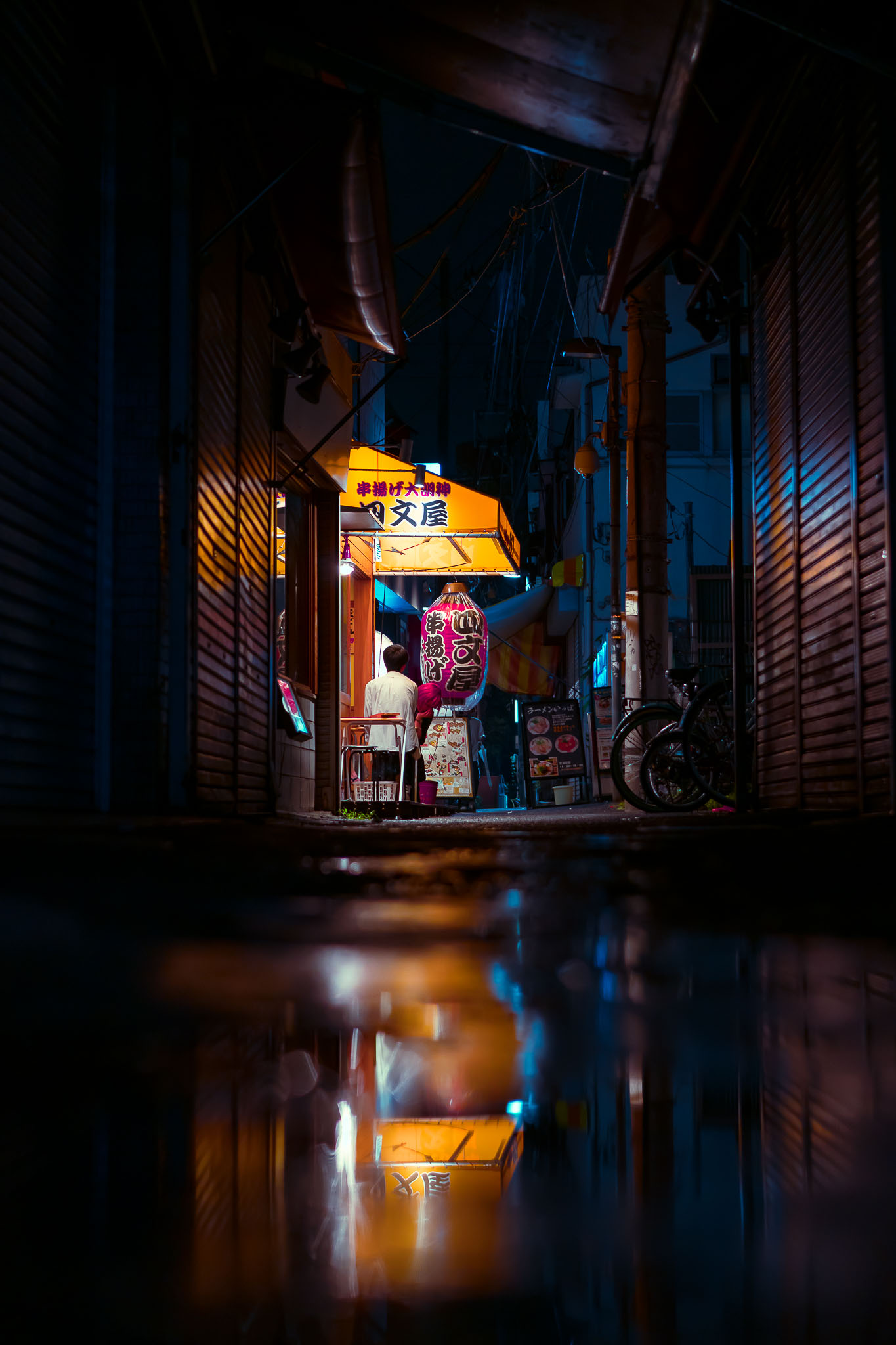 Narrow Koenji alley at night with a lit kushiage skewer restaurant sign and pink paper lantern reflected in a rain puddle