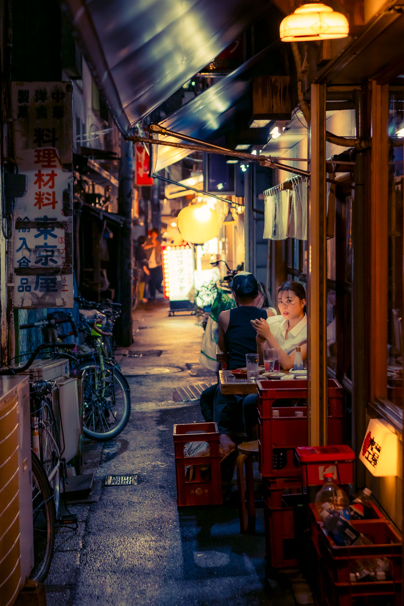 A couple seated at an outdoor table lit by a paper lantern in a narrow Koenji backstreet at night
