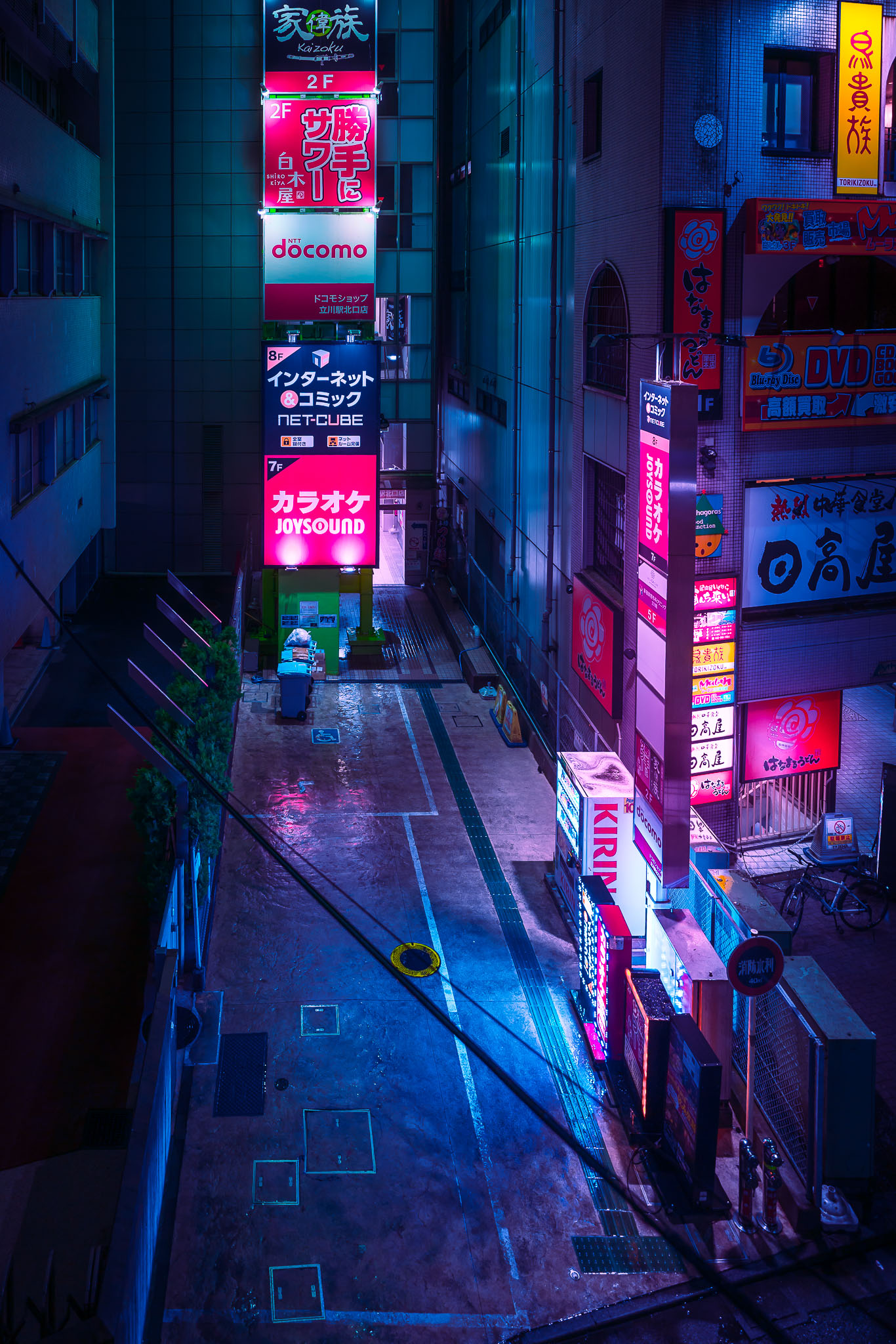 Backstreet in Tachikawa lit by a tall stacked sign for a docomo shop, JOYSOUND karaoke, and Net-Cube manga cafe, reflected on wet pavement