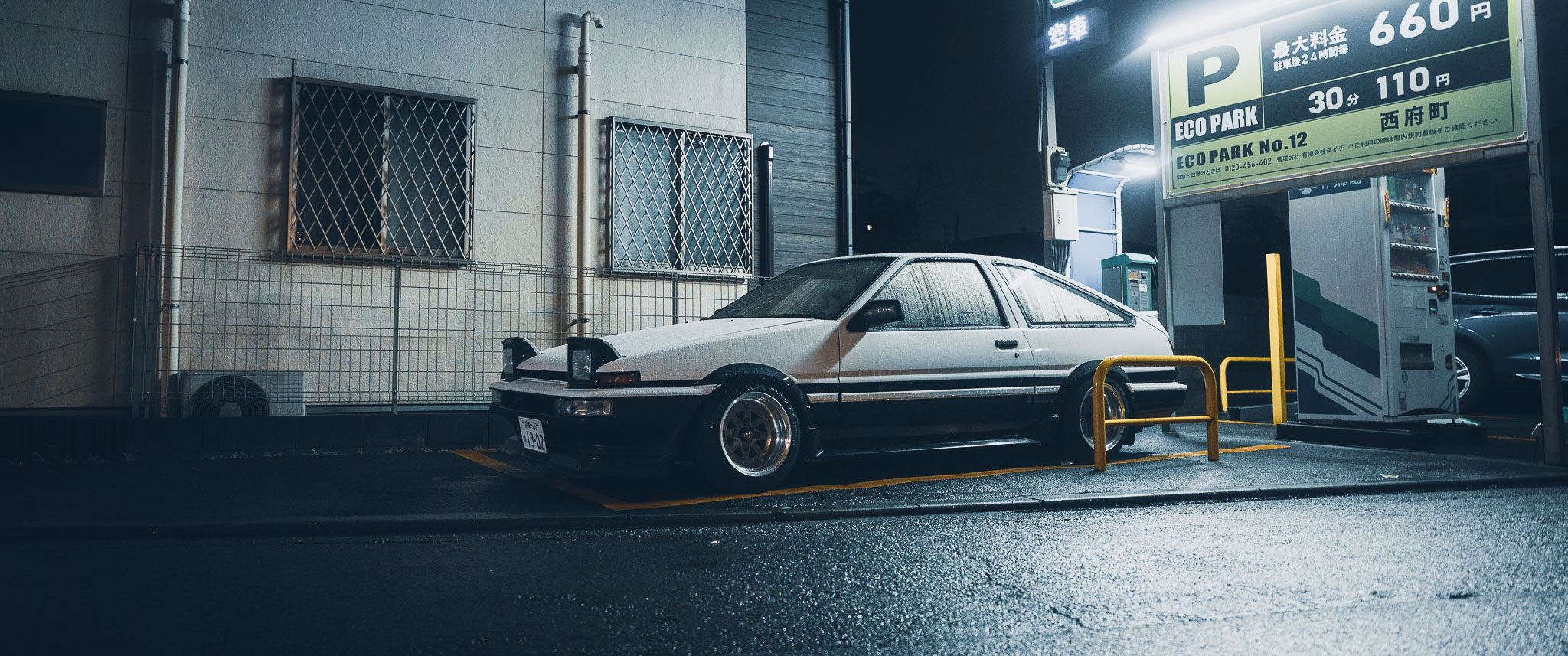 Toyota AE86 in a Japanese coin parking lot at night in the rain — full side view